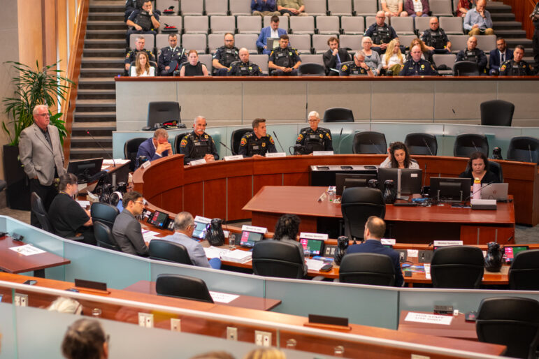 A wide, high-angle interior photograph shows a formal meeting underway inside a large, tiered municipal auditorium. Members of the board are seated along a curved wooden dais in the midground, accompanied by uniformed high-ranking police officers. In the foreground, additional staff members sit at long desks equipped with laptops and microphones. The background features steep, stadium-style seating populated by several uniform-clad police personnel and civilian observers beneath modern recessed lighting.