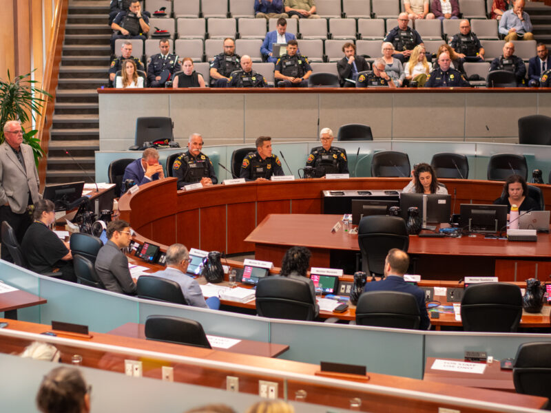 A wide, high-angle interior photograph shows a formal meeting underway inside a large, tiered municipal auditorium. Members of the board are seated along a curved wooden dais in the midground, accompanied by uniformed high-ranking police officers. In the foreground, additional staff members sit at long desks equipped with laptops and microphones. The background features steep, stadium-style seating populated by several uniform-clad police personnel and civilian observers beneath modern recessed lighting.