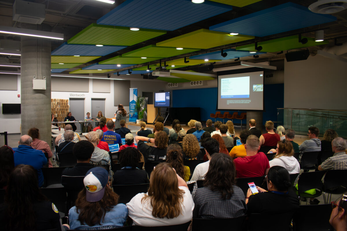 A wide, slightly elevated shot captures a densely seated audience attending a public meeting inside a large civic room. Dozens of attendees are viewed from behind, sitting in rows of black plastic chairs facing a raised presentation area. On the left, library board members are seated at tables facing the audience, while a delegate speaks at a podium. On the right, a large projection screen displays a slide presentation and a live video feed. The modern room features a thick concrete structural pillar, white walls, and a distinctive exposed ceiling fitted with large rectangular acoustic panels painted in vibrant blue and lime green.