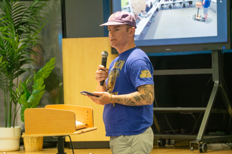 A medium shot captures a man speaking into a black microphone while holding a smartphone in his other hand during a public meeting. He is wearing a purple baseball cap, a blue t-shirt with Canadian Union of Postal Employees Local 548 branding, and light-coloured trousers, with visible tattoos on his left arm. He stands beside a small wooden podium. The background is softly out of focus, showing a large potted green plant, a light wood cabinet, and a large digital monitor displaying a live video feed of the meeting room layout under bright indoor lighting.