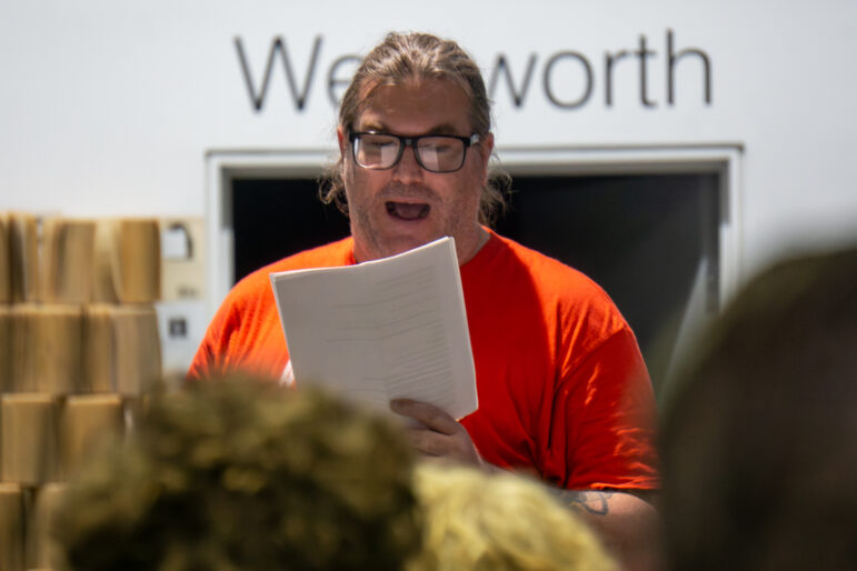 A close-up, eye-level photograph focuses on a man reading aloud from a multi-page printed document held in both hands during a public delegation. The man has long dark hair tied back, wears thick black rectangular glasses, and is dressed in a vibrant orange t-shirt. His mouth is open in mid-speech. The foreground is heavily blurred, showing the tops of the heads of seated audience members. The background, also softly out of focus, reveals a doorway topped with large black lettering spelling Wentworth, alongside a decorative wall installation constructed from stacked book pages.