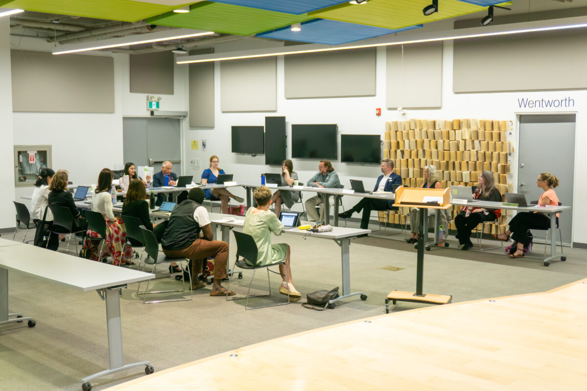 A wide shot captures a library board meeting where over a dozen participants sit along a U-shaped configuration of grey folding tables. Attendees, dressed in business-casual attire, interface with laptops and documents under bright, linear overhead fluorescent lights supplemented by acoustic ceiling panels in shades of green and blue. The background features white walls mounted with flat-screen monitors, an exit door, and a decorative partition wall composed of vertically stacked book pages next to a doorway labelled Wentworth.
