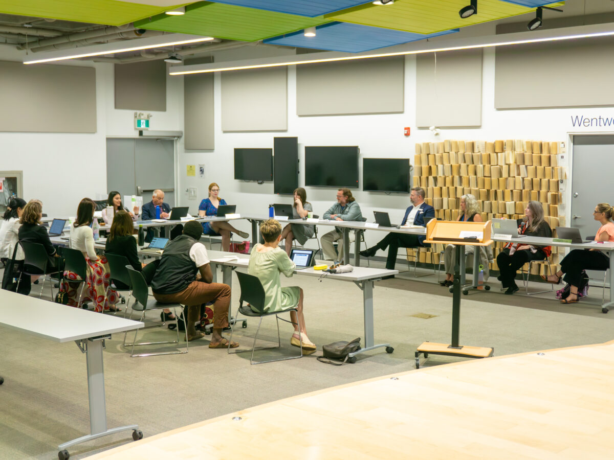 A wide shot captures a library board meeting where over a dozen participants sit along a U-shaped configuration of grey folding tables. Attendees, dressed in business-casual attire, interface with laptops and documents under bright, linear overhead fluorescent lights supplemented by acoustic ceiling panels in shades of green and blue. The background features white walls mounted with flat-screen monitors, an exit door, and a decorative partition wall composed of vertically stacked book pages next to a doorway labelled Wentworth.
