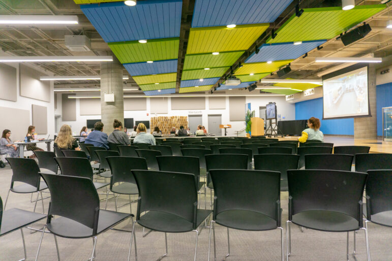 A wide, low-angle perspective looks across rows of empty black plastic chairs facing towards the library board members seated at the far end of the room. The expansive facility features an industrial ceiling accented by long rectangular panels of vibrant blue and lime green, a thick concrete supporting pillar, a wooden speaker podium, and a large projection screen displaying a live video feed on the right wall.
