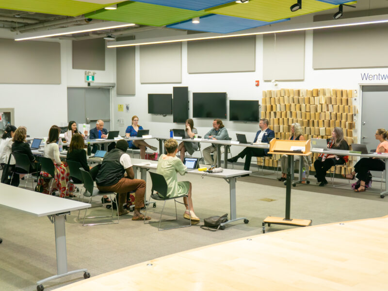 A wide shot captures a library board meeting where over a dozen participants sit along a U-shaped configuration of grey folding tables. Attendees, dressed in business-casual attire, interface with laptops and documents under bright, linear overhead fluorescent lights supplemented by acoustic ceiling panels in shades of green and blue. The background features white walls mounted with flat-screen monitors, an exit door, and a decorative partition wall composed of vertically stacked book pages next to a doorway labelled Wentworth.