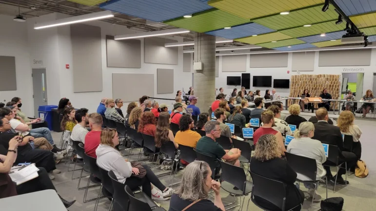 A wide-angle interior photograph taken from the side aisle of a large assembly space shows an audience seated in rows of black plastic chairs. Several chairs in the midground have blue printed signs reading "Reserved for Delegations" taped to their backrests. The attendees are facing towards the right side of the frame, where library board members are seated along tables near a doorway labelled Wentworth. The modern room features a large cylindrical concrete structural pillar, white walls, and a distinctive exposed ceiling fitted with rectangular acoustic panels painted in bright blue and lime green.
