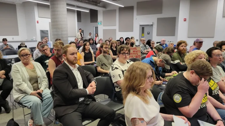 A medium close-up photograph focuses on a densely packed crowd of meeting attendees seated in black chairs, all looking forward toward an unseen speaker. In the front left, a man in a dark suit and white collared shirt sits with his hands clasped. On the right, an individual wears a black t-shirt featuring a yellow circular logo for CUPE Local 932. Further back in the crowd, a person holds up a handmade cardboard sign with red hand-painted text reading "HOARDING MAKES LIBRARIES LESS SAFE!". The room is brightly illuminated with flat indoor lighting, revealing acoustic wall panels and a concrete structural pillar in the background.