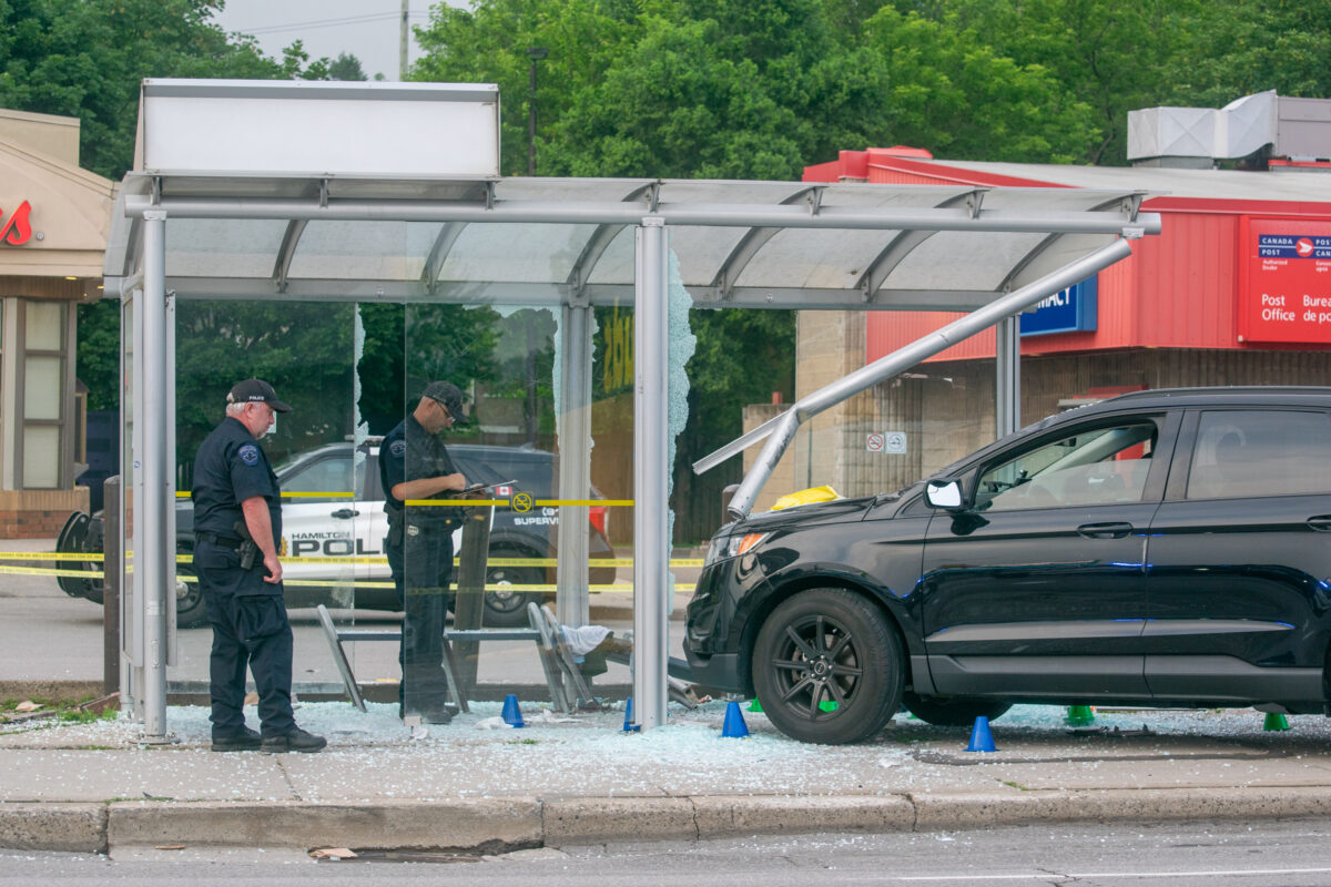 A daytime medium shot captures two Hamilton Police Service officers standing amidst the mangled structural framing of a public transit shelter recently struck by a black SUV. The front end of the vehicle rests entirely on the pedestrian walkway, having destroyed the right-side support pillars of the shelter and caused the curved roof frame to buckle downward onto the hood. The sidewalk is completely covered in shattered safety glass interspersed with blue-coloured evidence markers. One officer wearing a dark uniform and a police cap takes notes on a clipboard, while a second officer observes the immediate wreckage. Behind the officers, a marked Hamilton Police cruiser is parked alongside yellow police tape, with a red Canada Post outlet and other commercial retail storefronts visible in the background under flat, overcast daylight conditions.