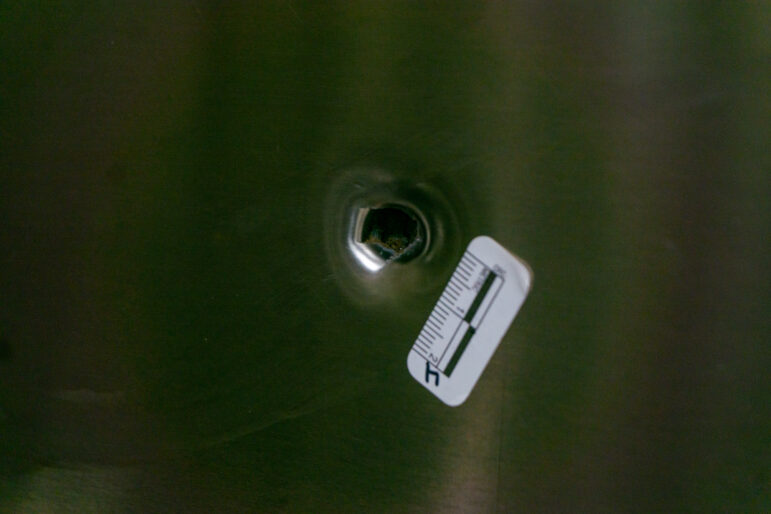 A macro close-up photograph captures a single bullet hole piercing a smooth, brushed metal surface. The metal surrounding the puncture is dented inwards from the impact, revealing jagged, dark edges within the central cavity. Positioned immediately to the lower right of the impact point is a small, white adhesive police evidence scale featuring black metric ruler markings and a handwritten number four. The lighting is dim and diffused, creating soft vertical reflections across the dark, textured metallic background while highlighting the structural damage to the panel.