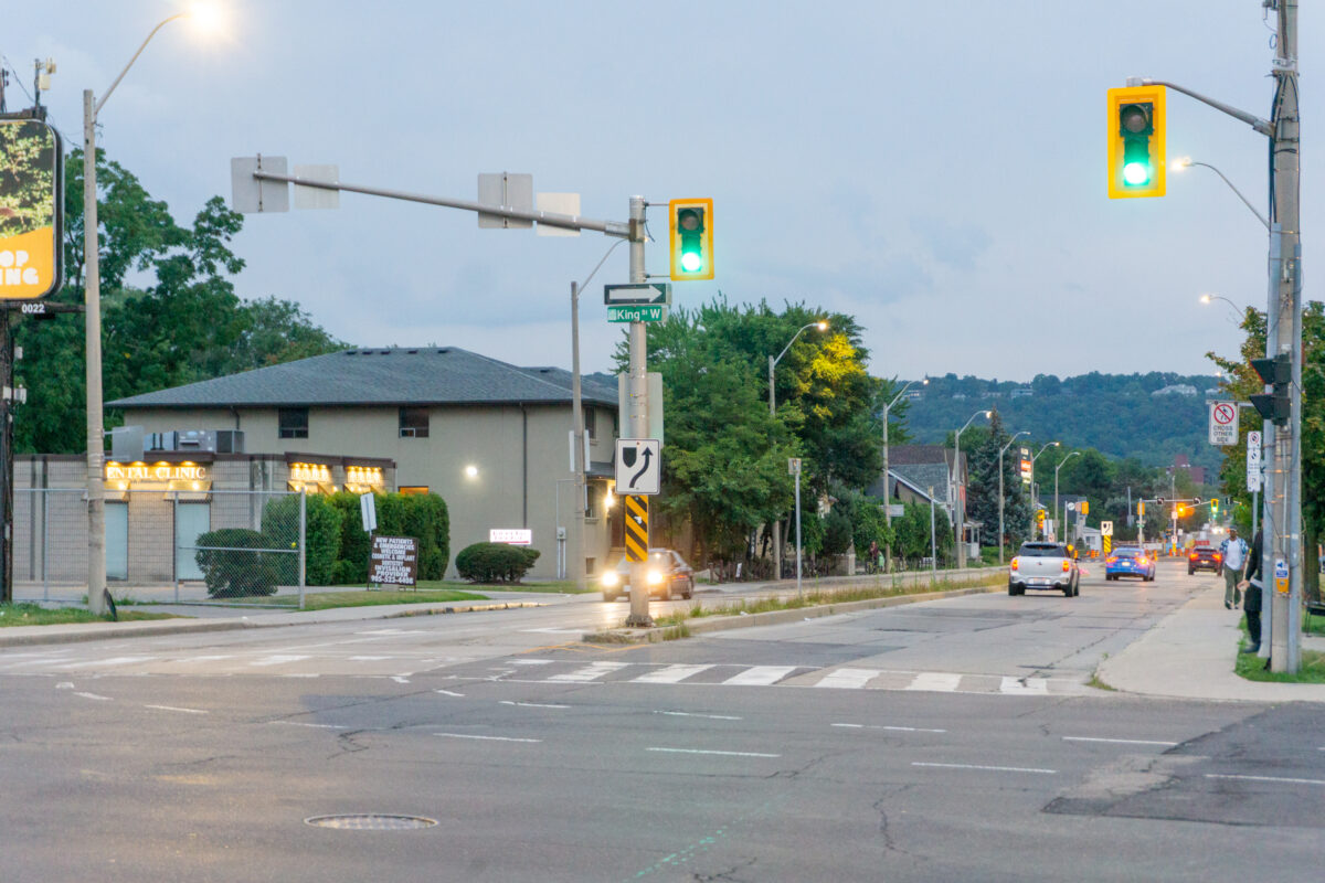 A street-level perspective looks south down Dundurn Street South from its intersection with King Street West. Active green traffic signals suspended from metal mast arms illuminate the foreground above a marked pedestrian crosswalk on the asphalt road surface. To the left, a single-storey commercial building housing a dental clinic stands behind a chain-link fence and manicured bushes, while vehicles travel along the multi-lane roadway. To the right, pedestrians walk along the concrete sidewalk adjacent to wooden utility poles. Lush green trees line the street, leading toward the elevated silhouette of the Niagara Escarpment under a soft, overcast twilight sky.