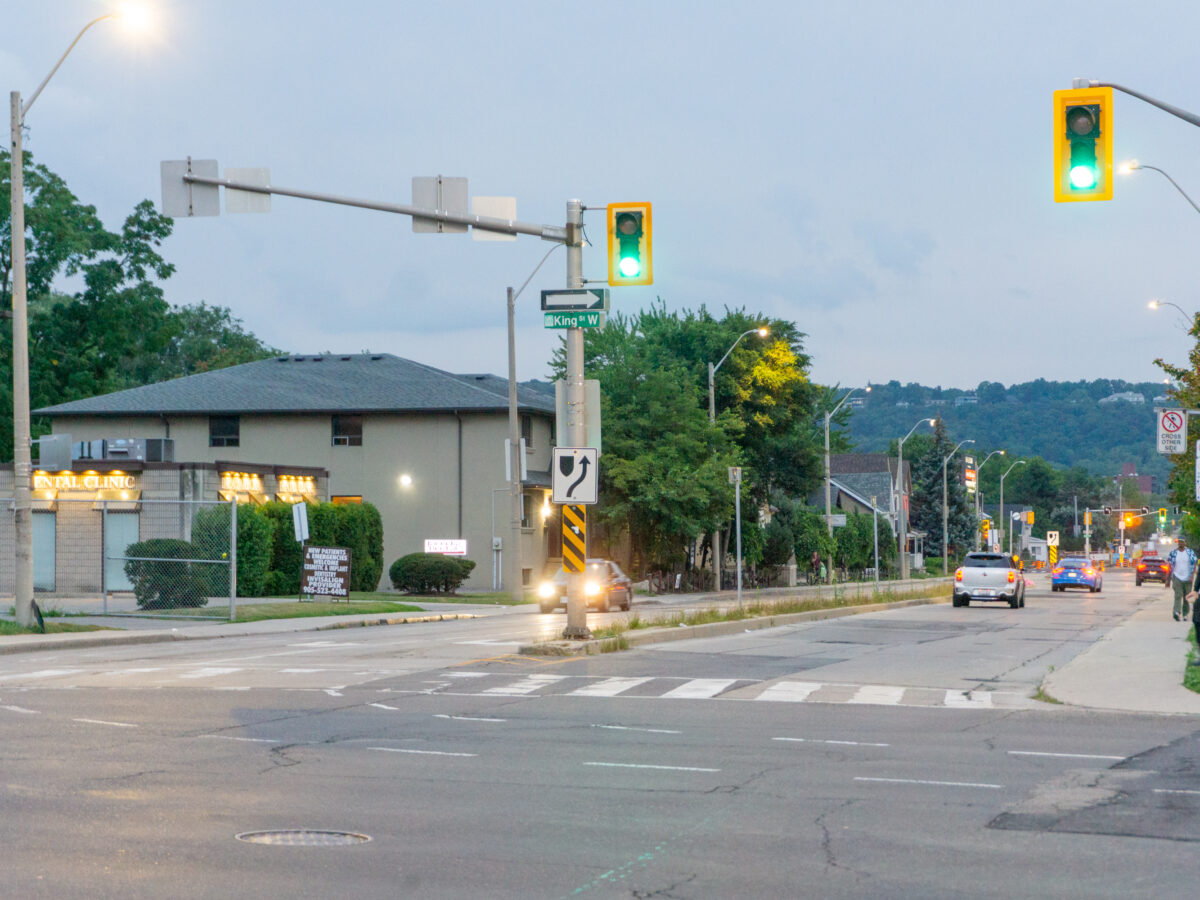 A street-level perspective looks south down Dundurn Street South from its intersection with King Street West. Active green traffic signals suspended from metal mast arms illuminate the foreground above a marked pedestrian crosswalk on the asphalt road surface. To the left, a single-storey commercial building housing a dental clinic stands behind a chain-link fence and manicured bushes, while vehicles travel along the multi-lane roadway. To the right, pedestrians walk along the concrete sidewalk adjacent to wooden utility poles. Lush green trees line the street, leading toward the elevated silhouette of the Niagara Escarpment under a soft, overcast twilight sky.