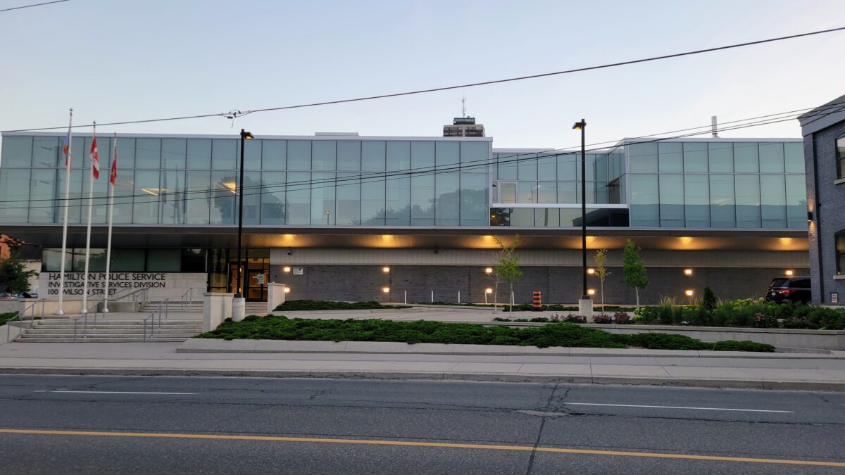 A wide, eye-level exterior photograph showcases the modern architectural facade of the Hamilton Police Service Investigative Services Division building at 100 Wilson Street during twilight. The structure features a prominent, cantilevered upper storey enclosed in an extensive curtain wall of green-tinted reflective glass panels, set above a ground level clad in dark grey brick and concrete masonry. Recessed amber lighting fixtures cast downward glows across the lower exterior walls and along the underside of the upper overhang. In the foreground, an asphalt roadway marked with yellow centre lines borders a concrete sidewalk and terraced planting beds filled with low evergreen shrubs and young trees. Wide concrete steps and an accessible ramp lead to the main entrance on the left, flanked by three tall metal flagpoles flying Canadian, provincial, and service flags. Overhead utility lines stretch horizontally across the pale dusk sky.