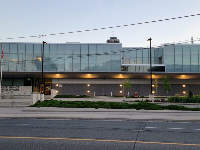 A wide, eye-level exterior photograph showcases the modern architectural facade of the Hamilton Police Service Investigative Services Division building at 100 Wilson Street during twilight. The structure features a prominent, cantilevered upper storey enclosed in an extensive curtain wall of green-tinted reflective glass panels, set above a ground level clad in dark grey brick and concrete masonry. Recessed amber lighting fixtures cast downward glows across the lower exterior walls and along the underside of the upper overhang. In the foreground, an asphalt roadway marked with yellow centre lines borders a concrete sidewalk and terraced planting beds filled with low evergreen shrubs and young trees. Wide concrete steps and an accessible ramp lead to the main entrance on the left, flanked by three tall metal flagpoles flying Canadian, provincial, and service flags. Overhead utility lines stretch horizontally across the pale dusk sky.