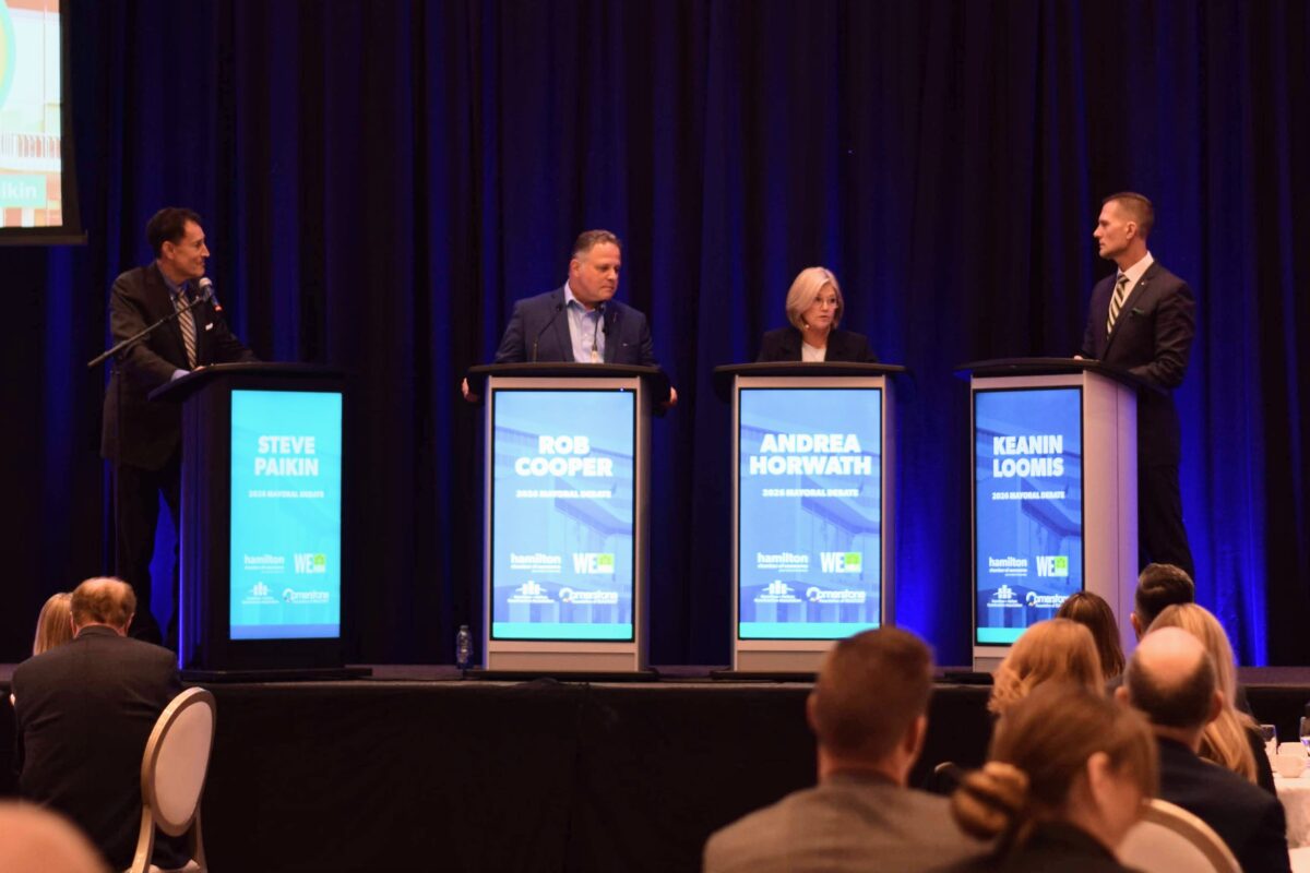 A wide, eye-level photograph captures four participants standing at individual lecterns on an elevated stage during an indoor civic debate, viewed from behind a seated audience. From left to right, four black podiums feature illuminated digital displays with bright cyan-blue panels that display the names Steve Paikin, Rob Cooper, Andrea Horwath, and Keanin Loomis beneath the label 2026 Mayoral Debate alongside institutional sponsor logos. Moderator Steve Paikin stands at the far left lectern wearing a dark suit and striped tie, speaking into a gooseneck microphone. Candidate Rob Cooper stands at the second lectern in a dark blazer and collared dress shirt. Incumbent Mayor Andrea Horwath stands at the third podium dressed in a dark suit jacket, looking attentively across the stage. Candidate Keanin Loomis occupies the fourth lectern on the right in a dark tailored suit and patterned tie. A wall-to-wall backdrop of pleated dark blue stage curtains hangs behind the speakers, highlighted by directional overhead stage spotlights, while the silhouettes of attendees seated at banquet tables frame the lower foreground under ambient event lighting.