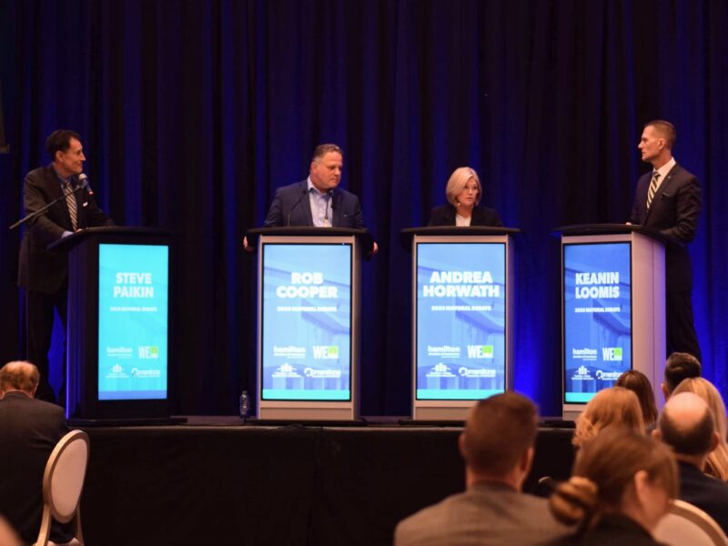 A wide, eye-level photograph captures four participants standing at individual lecterns on an elevated stage during an indoor civic debate, viewed from behind a seated audience. From left to right, four black podiums feature illuminated digital displays with bright cyan-blue panels that display the names Steve Paikin, Rob Cooper, Andrea Horwath, and Keanin Loomis beneath the label 2026 Mayoral Debate alongside institutional sponsor logos. Moderator Steve Paikin stands at the far left lectern wearing a dark suit and striped tie, speaking into a gooseneck microphone. Candidate Rob Cooper stands at the second lectern in a dark blazer and collared dress shirt. Incumbent Mayor Andrea Horwath stands at the third podium dressed in a dark suit jacket, looking attentively across the stage. Candidate Keanin Loomis occupies the fourth lectern on the right in a dark tailored suit and patterned tie. A wall-to-wall backdrop of pleated dark blue stage curtains hangs behind the speakers, highlighted by directional overhead stage spotlights, while the silhouettes of attendees seated at banquet tables frame the lower foreground under ambient event lighting.