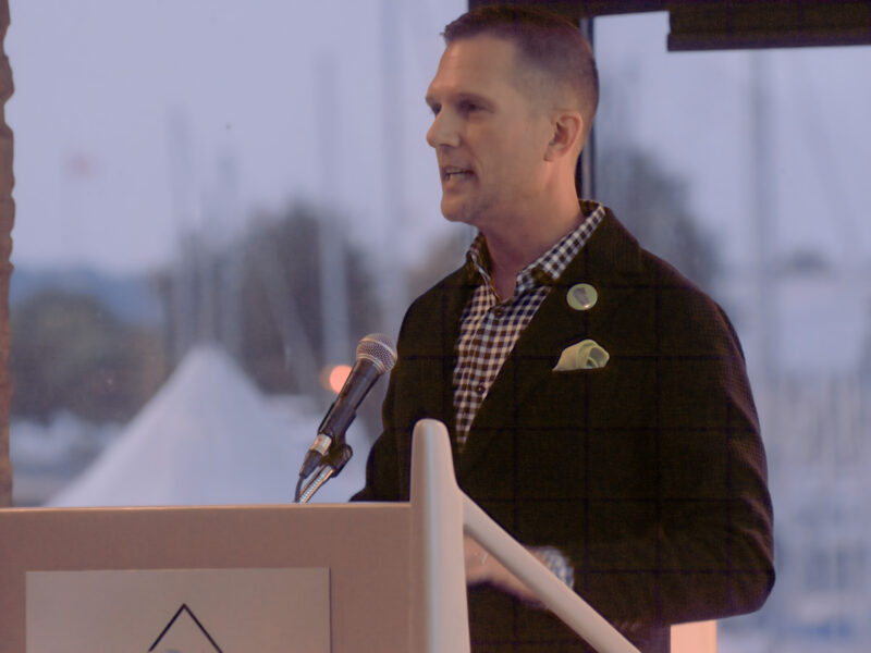 A medium close-up photograph captures Keanin Loomis in profile speaking from behind a light-coloured wooden lectern. He is dressed in a dark textured blazer over a black-and-white gingham patterned collared shirt, complemented by a folded light pocket square and a small circular lapel pin. A black microphone mounted on a stand extends toward him in the foreground. In the background, expansive floor-to-ceiling windows look out onto a waterfront scene at dusk, showing the soft silhouettes of boats and a white outdoor event marquee under warm, subdued indoor lighting.