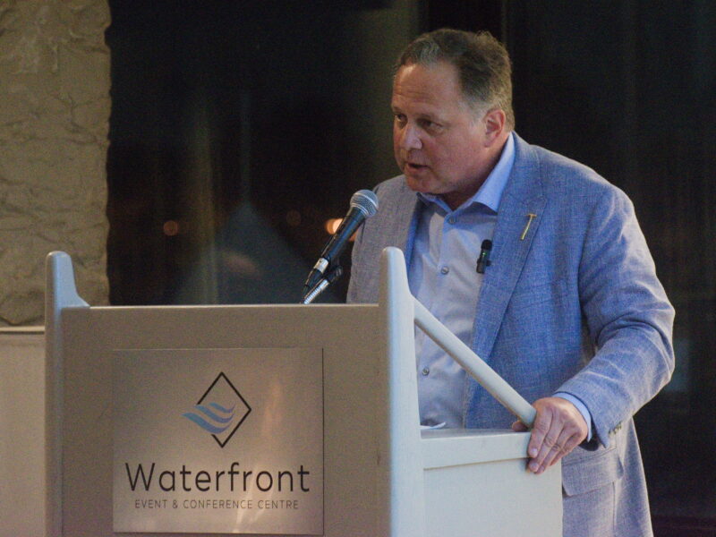 A medium shot shows Rob Cooper speaking to an audience from behind a light grey lectern mounted with a metallic logo plaque reading Waterfront Event and Conference Centre. He wears a textured blue sport coat over an unbuttoned light blue dress shirt accented with a gold lapel pin and a small clipped microphone. Leaning forward slightly, he rests both hands on the top corners of the podium while addressing a stand-mounted microphone. The background consists of dark window glass reflecting faint outdoor lights against a textured stone architectural column on the left under soft indoor spotlighting.